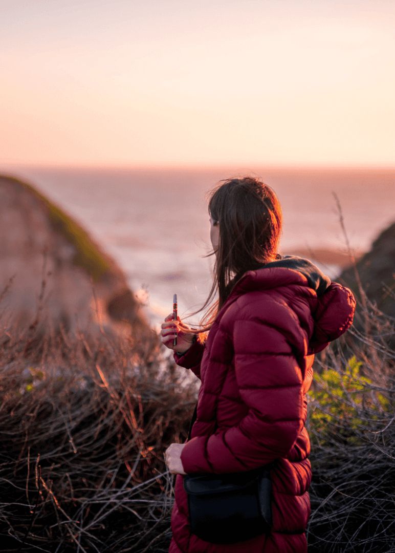 A woman wearing a red jacket holding her weed vape pen from Highway Cannabis Dispensary while standing on a cliff overlooking one of LA's beaches at sunset.